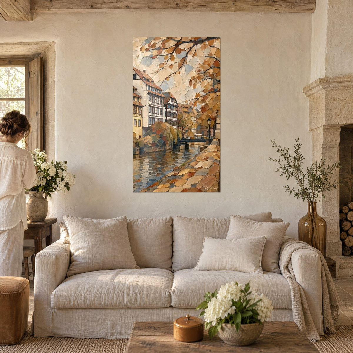 Gemälde mit beigen und ockerfarbenen Fachwerkhäusern unter mosaikartigem Laubwerk, Kanal und Mäuerchen, über einem Sofa in einem hellen Wohnzimmer