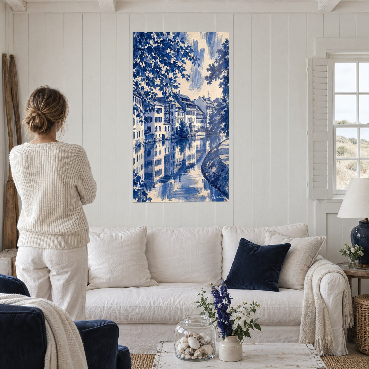 Cuadro canal con casas de entramado de madera, azul cobalto y crema, reflejos en el agua, encima de un sofá en un salón luminoso