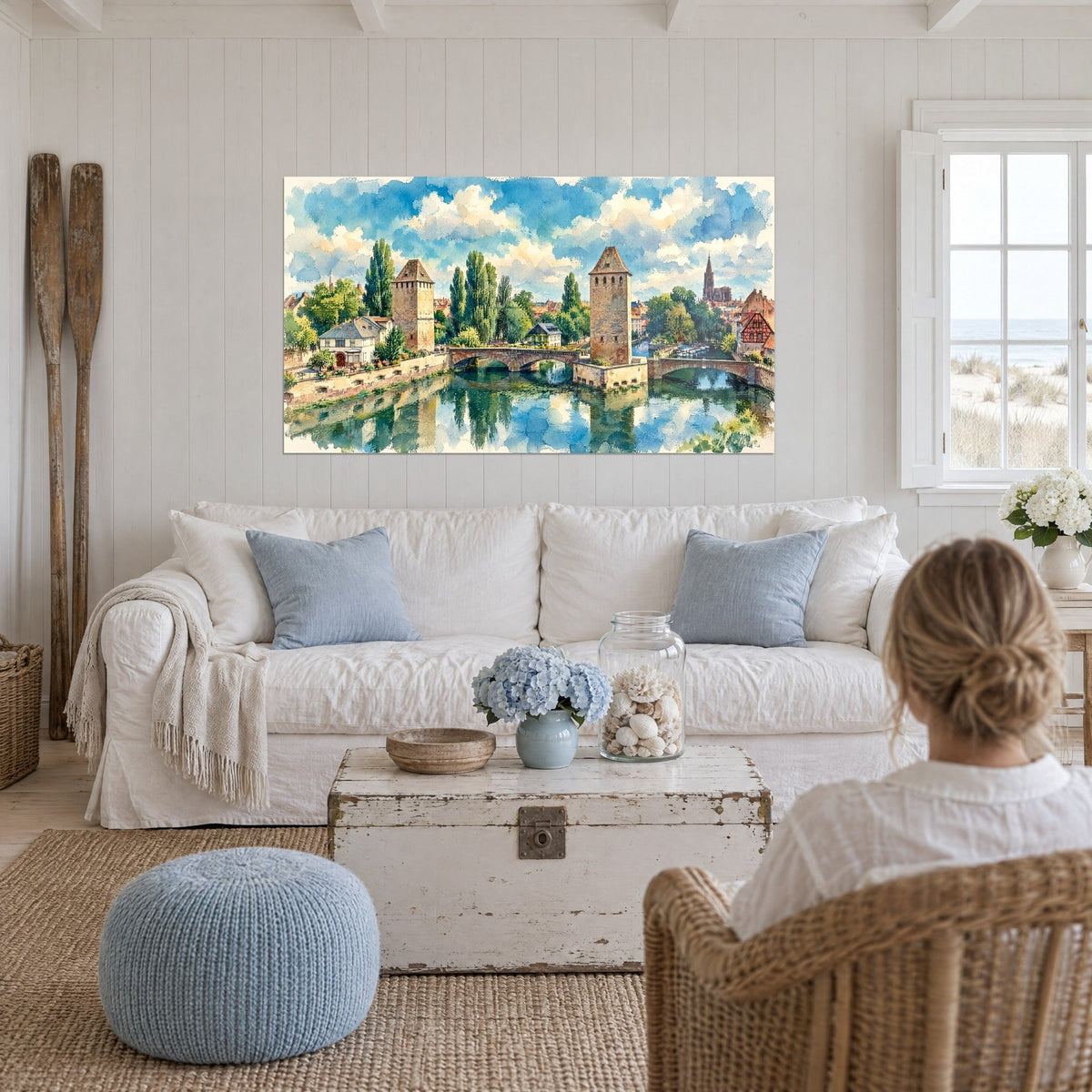 Schilderij Straatsburg in aquarel, beige torens en smaragdgroen gebladerte, vrouw van achteren, boven een bank in een lichte woonkamer