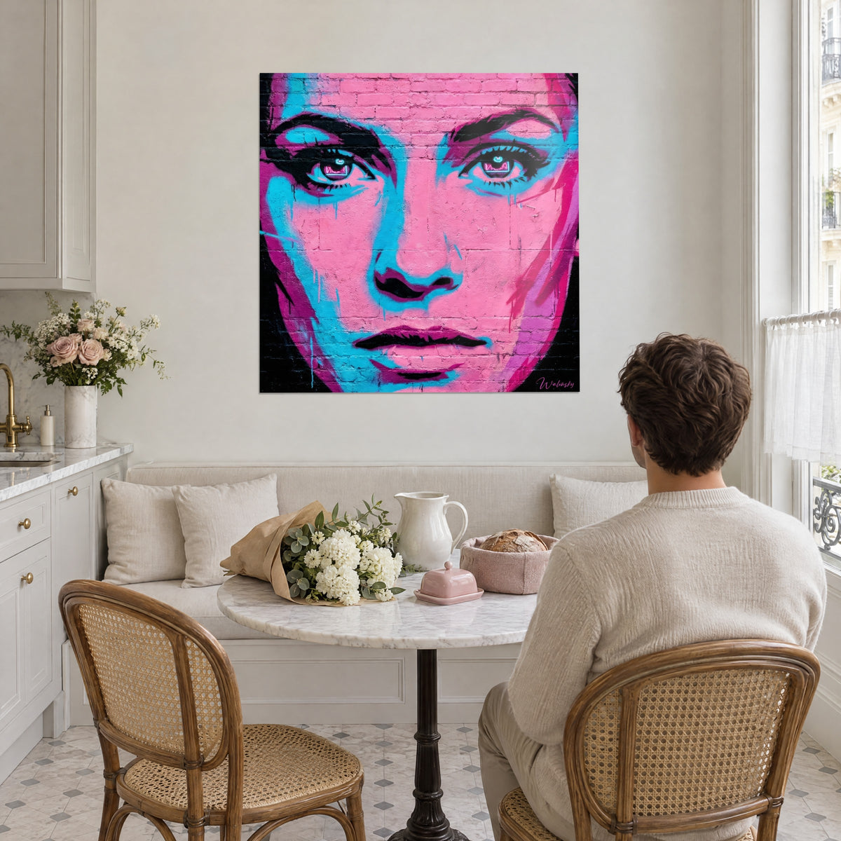 Cuadro rostro de grafiti de ojos eléctricos, rosa chicle, turquesa y negro, colgado detrás de un hombre de espaldas sentado a una mesa