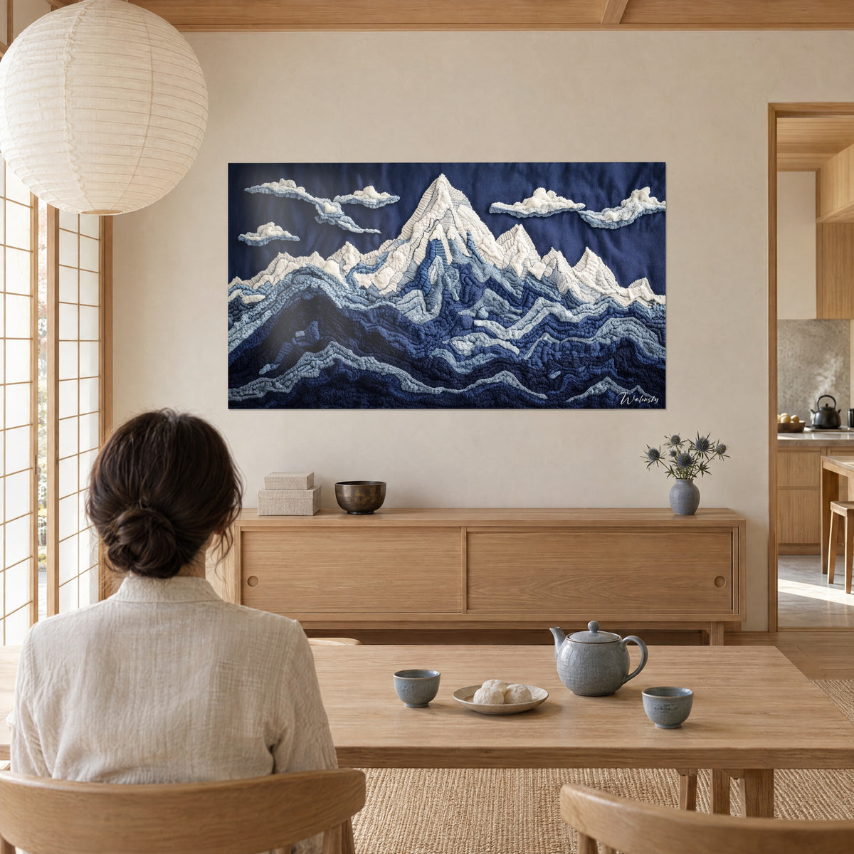Cuadro montaña en hilos tejidos, cima blanco crema, laderas azul glaciar y marino, contemplado por una mujer de espaldas a la mesa