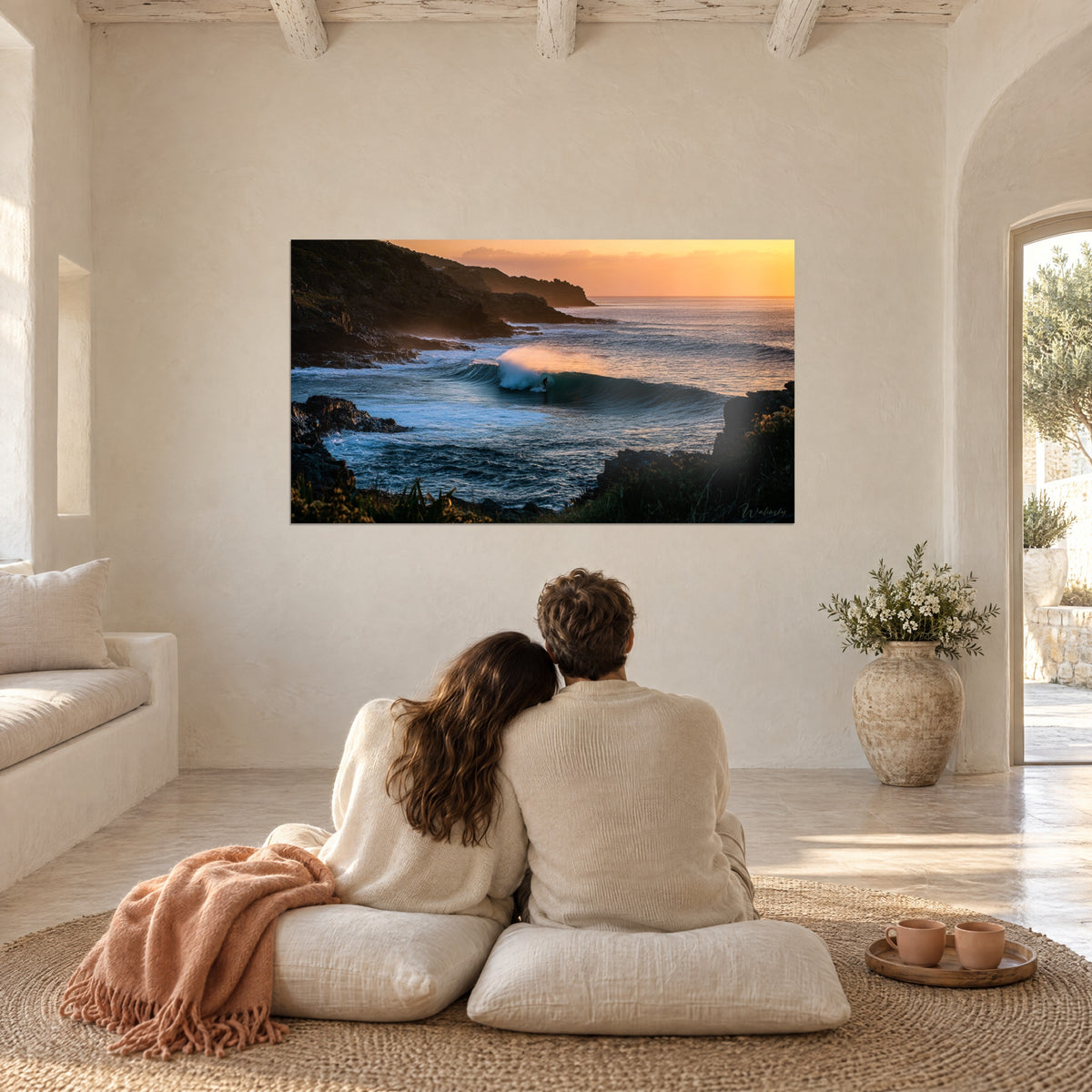 Tableau côte sauvage et vague dorée, roches brunes et eau indigo, regardé par un couple de dos assis sur des coussins crème