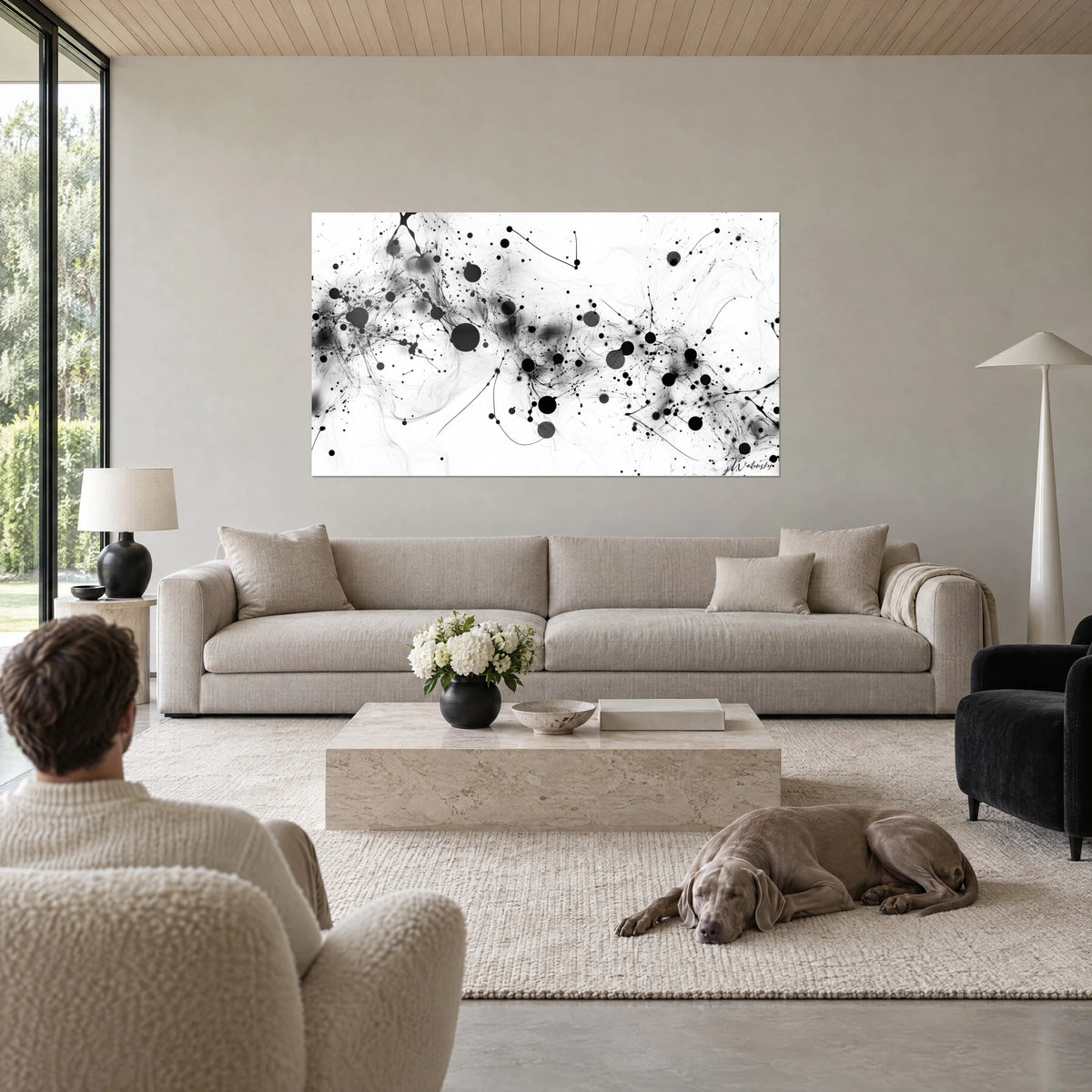 Tableau constellation d'encre noire et grise éclaboussant un fond blanc, observée par un Weimaraner couché, au-dessus d'un canapé dans un salon lumineux