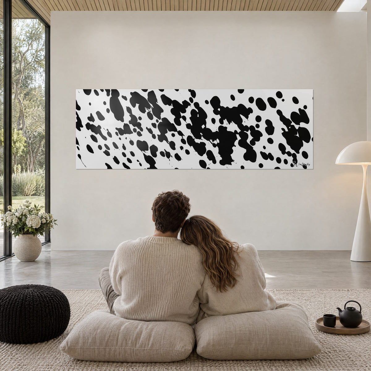 Cuadro con proyecciones de tinta negra sobre blanco brillante, pared crema, pareja abrazada sobre cojines beige frente al ventanal