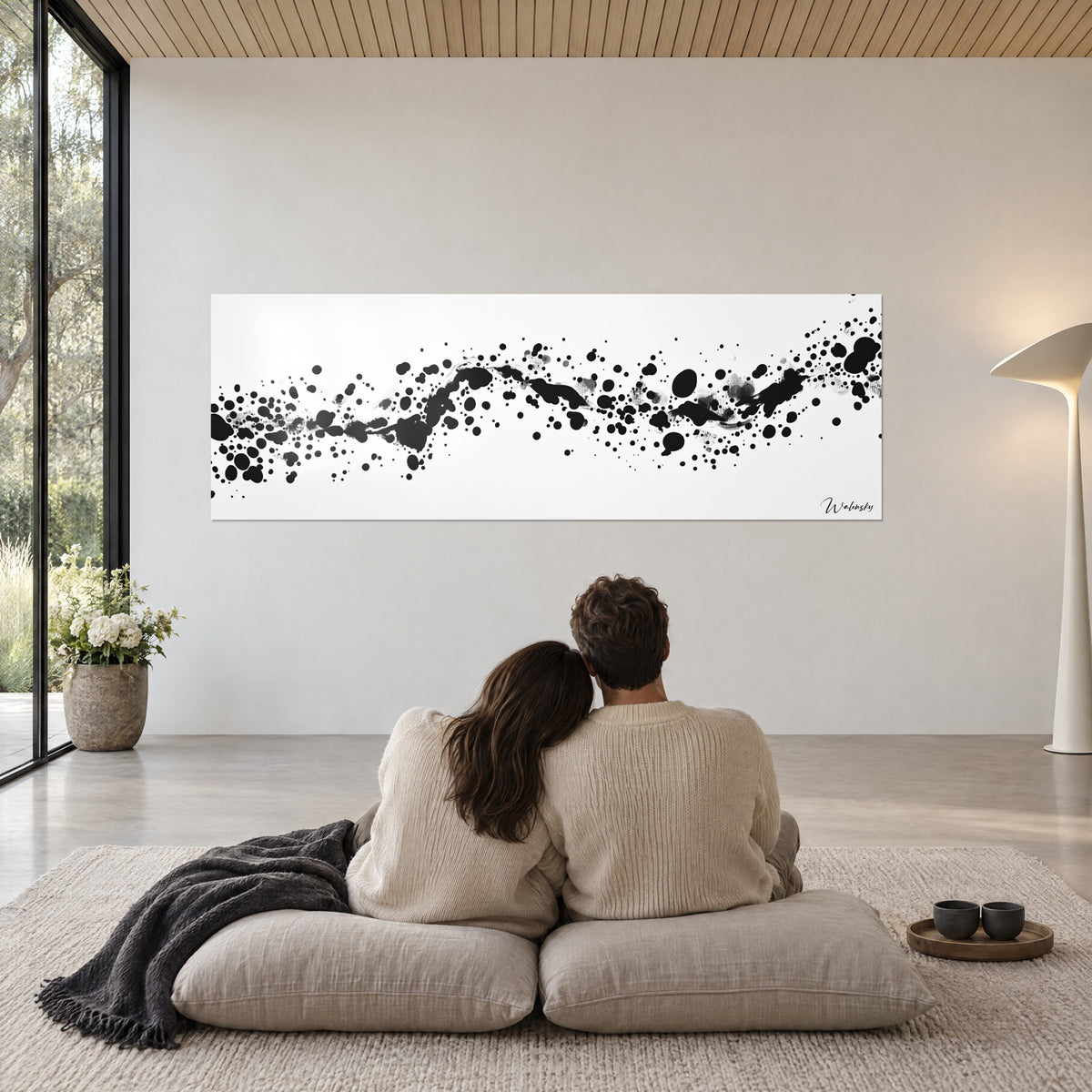 Cuadro de salpicaduras de tinta azabache en cinta sinuosa sobre fondo blanco, pareja abrazada de espaldas sobre cojines beige, manta antracita