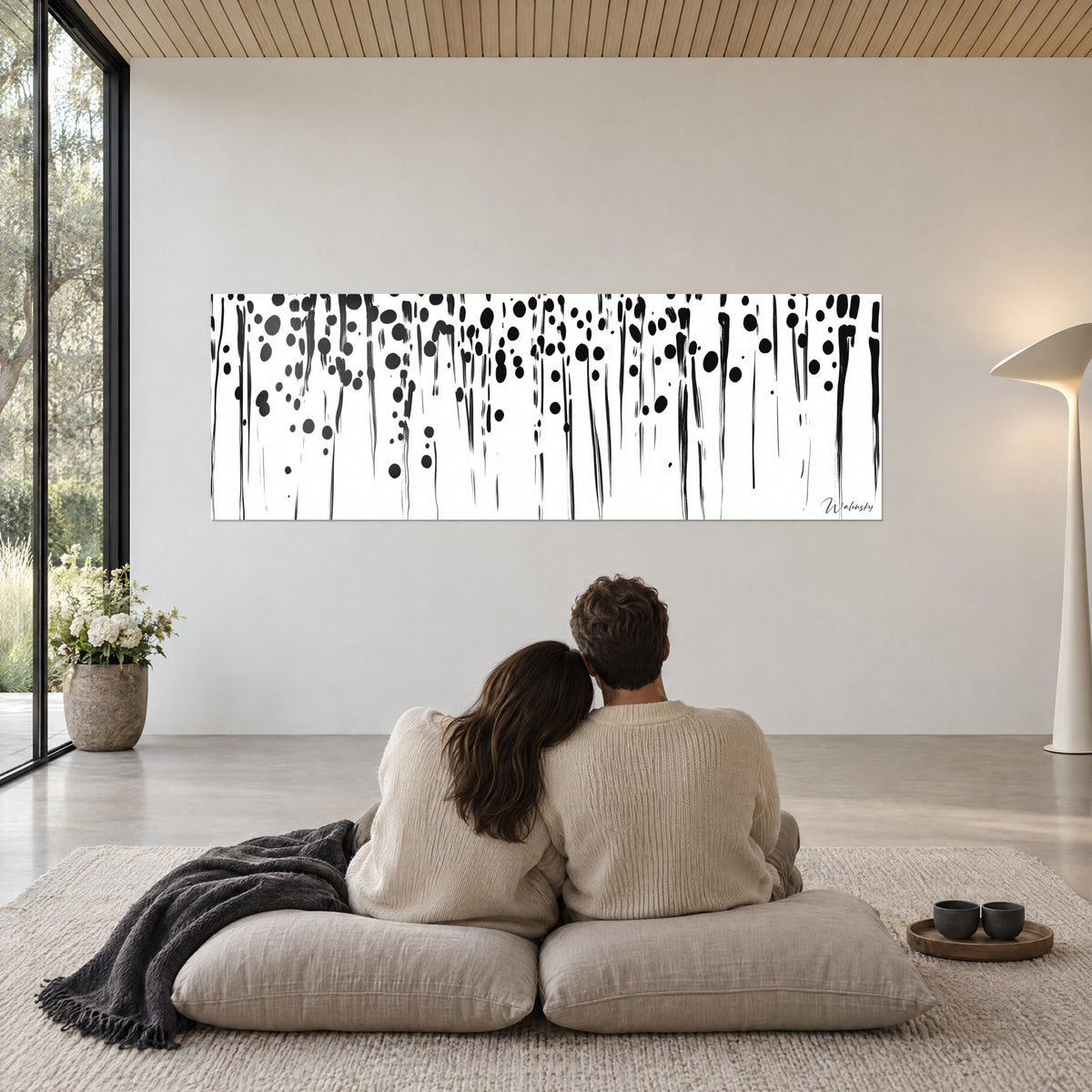 Quadro di macchie nere sparse e tratti verticali su bianco, coppia seduta di spalle su cuscini beige, soggiorno luminoso