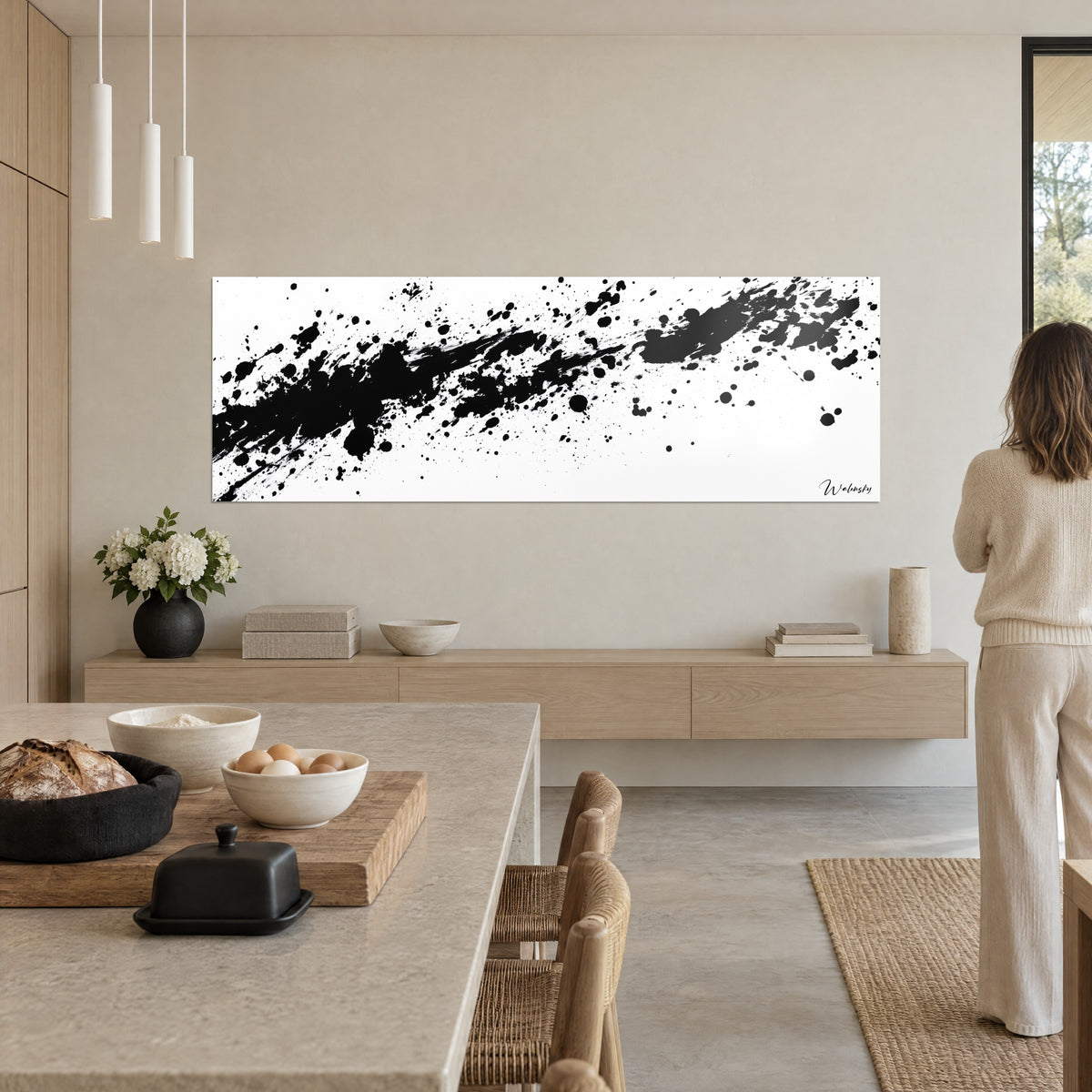Tableau jet de peinture noire en diagonale sur fond blanc crème, femme de dos près d'une cuisine ouverte en bois clair