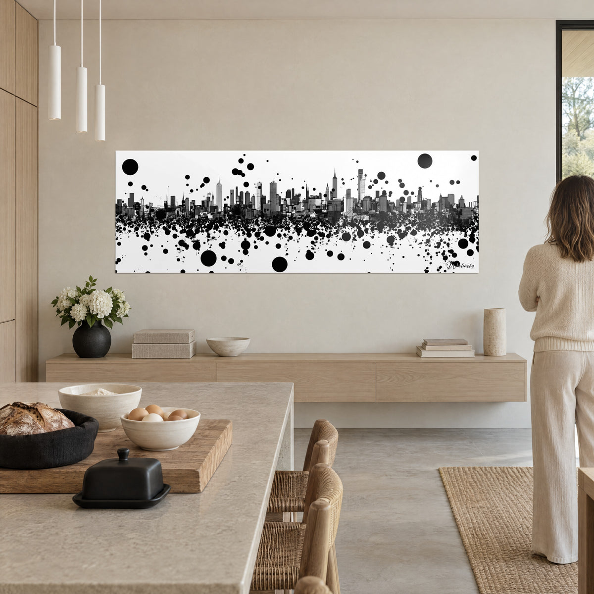 Quadro grattacieli disegnati a tratto nero, macchie d'inchiostro sparse su fondo bianco, donna di spalle in una cucina aperta beige