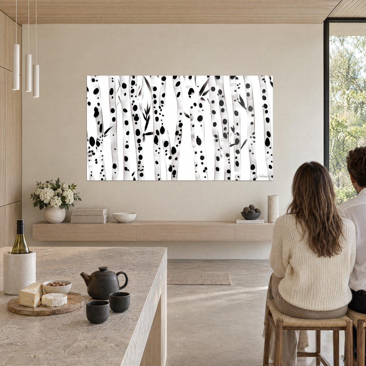 Cuadro de cañas esbeltas con puntos de tinta negra sobre fondo blanco, colgado en la pared de una cocina abierta de madera clara, pareja sentada a la mesa