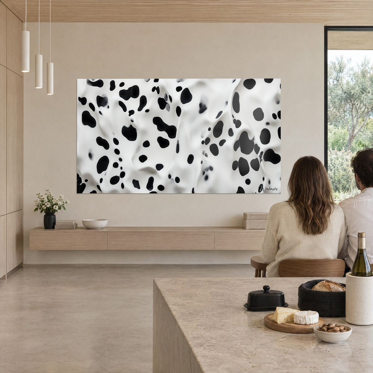 Tableau peau de vache stylisée, éclaboussures noires sur blanc satiné, deux personnes de dos devant un îlot de cuisine beige dans un séjour ouvert