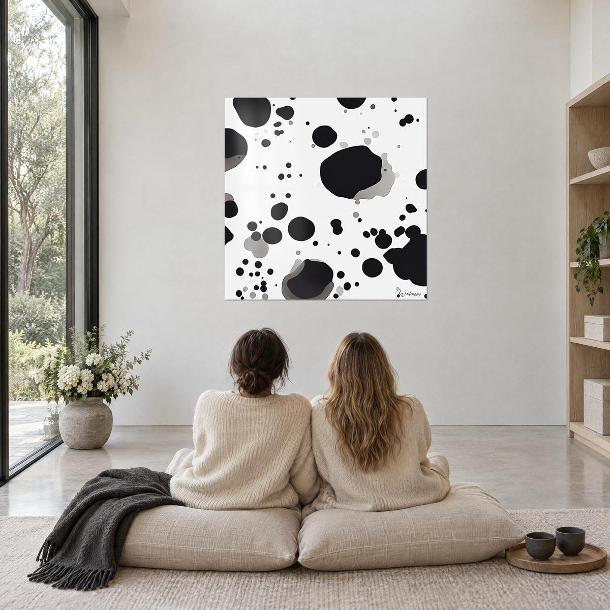 Tableau mosaïque de macules noires et grises sur fond ivoire, contemplé par deux femmes de dos assises sur des coussins beiges