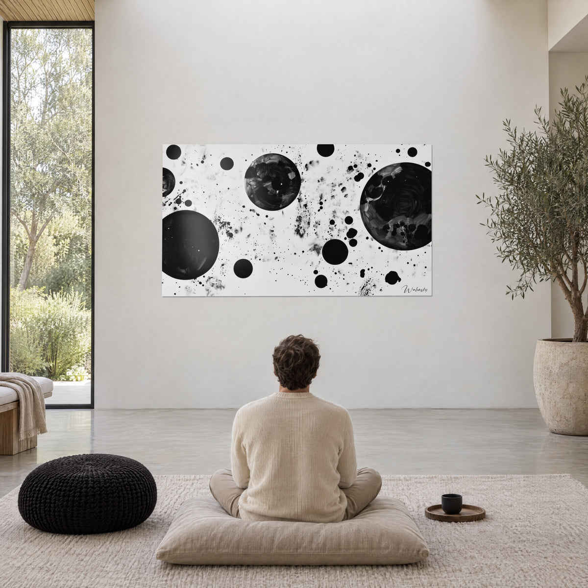 Cuadro lunas de tinta sobre fondo blanco, destellos negros y grises, frente a un hombre sentado en meditación sobre un cojín beige