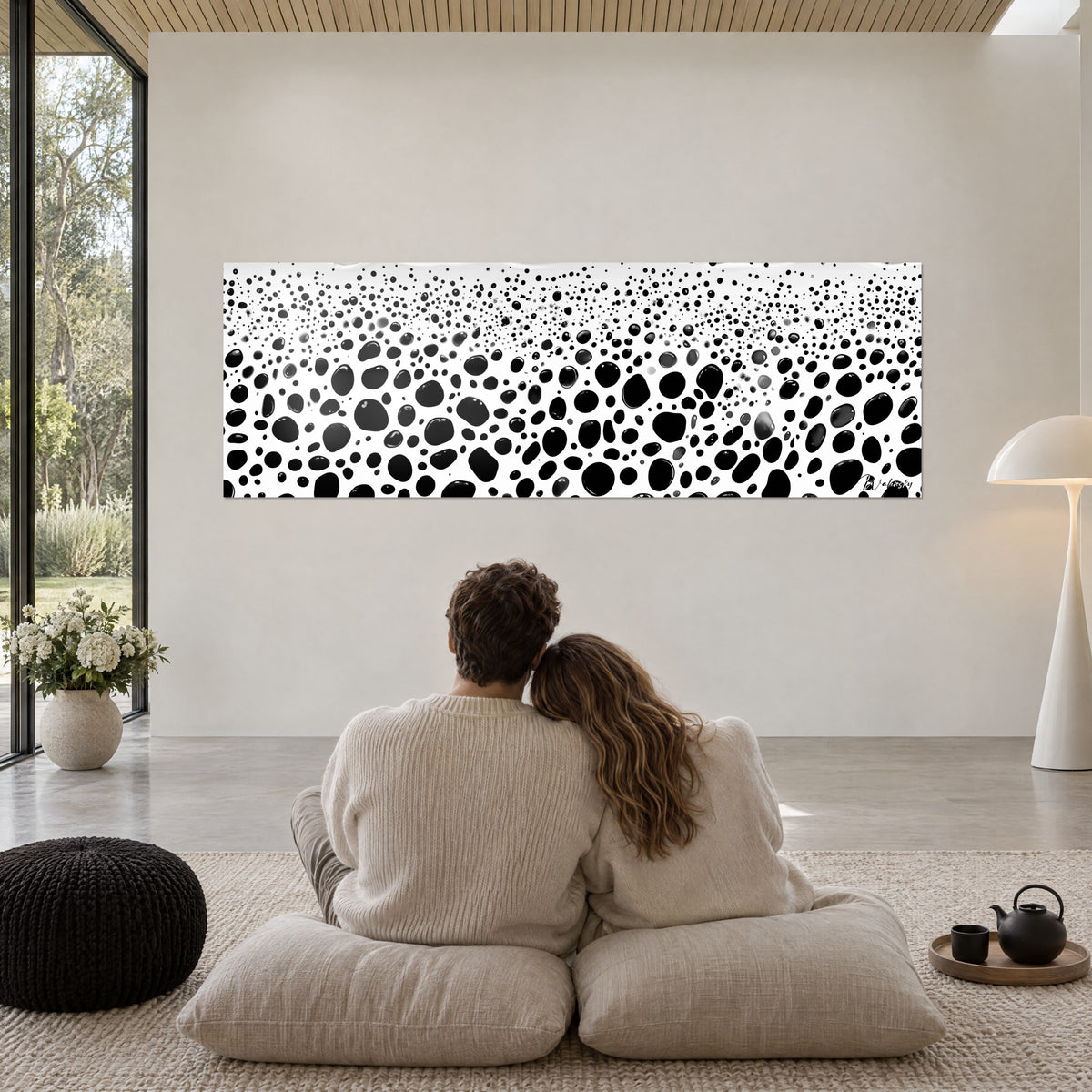 Cuadro con una constelación de burbujas negras brillantes sobre blanco, dos personas de espaldas abrazadas sobre cojines frente a una pared beige
