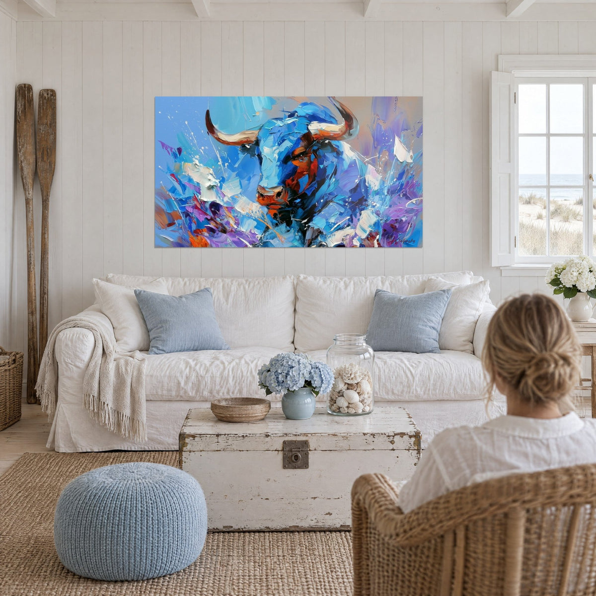 Cuadro Longhorn azul celeste con cuernos marfil y hocico rojizo, destellos violetas, mujer de espaldas sobre un sofá en un salón luminoso