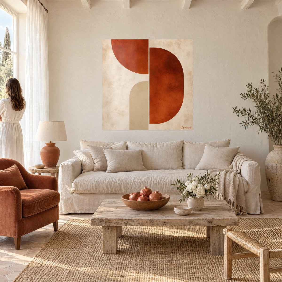 Wall art with rust half-circles and a beige arch on a cream background, woman in a white dress seen from behind, above a sofa in a bright living room