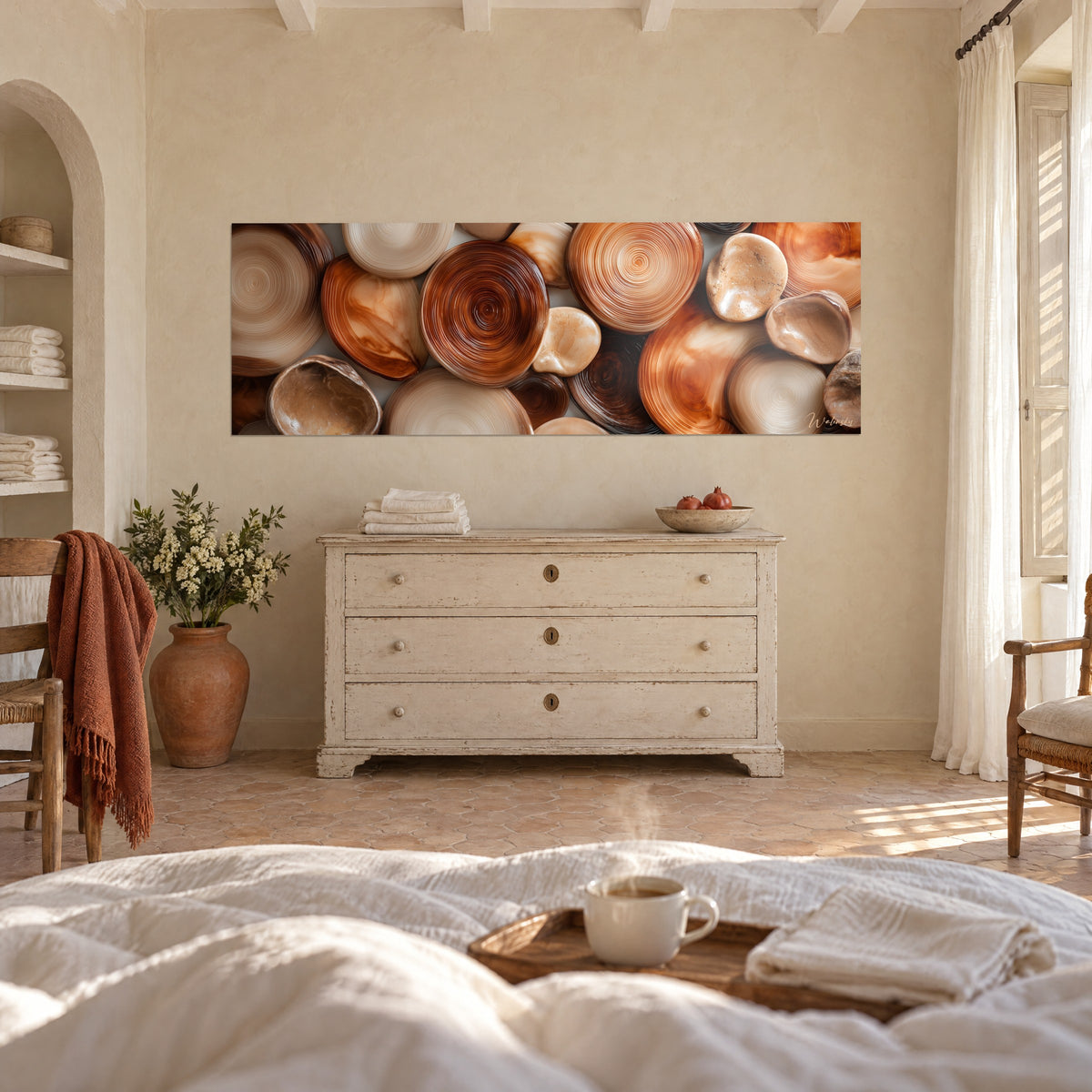 Quadro di ciottoli dagli anelli stretti, caramello e marrone cioccolato, di fronte a una cassettiera bianca, sopra un letto in una camera luminosa
