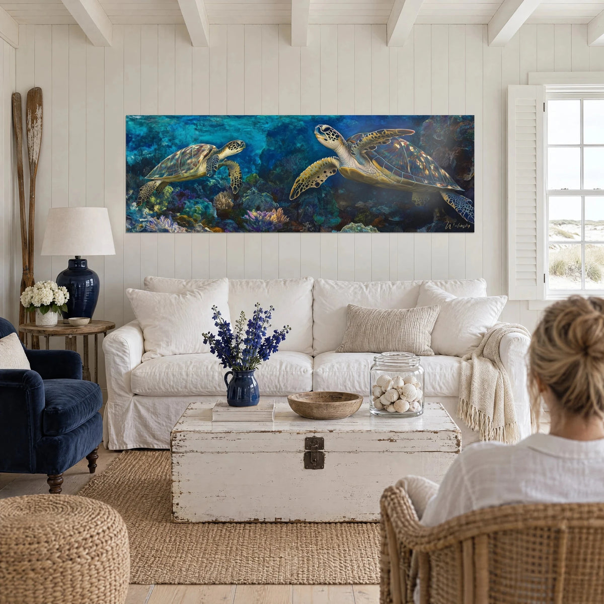 Wall art sea turtle gliding over a turquoise and gold reef, two figures seen from behind, above a sofa in a bright living room