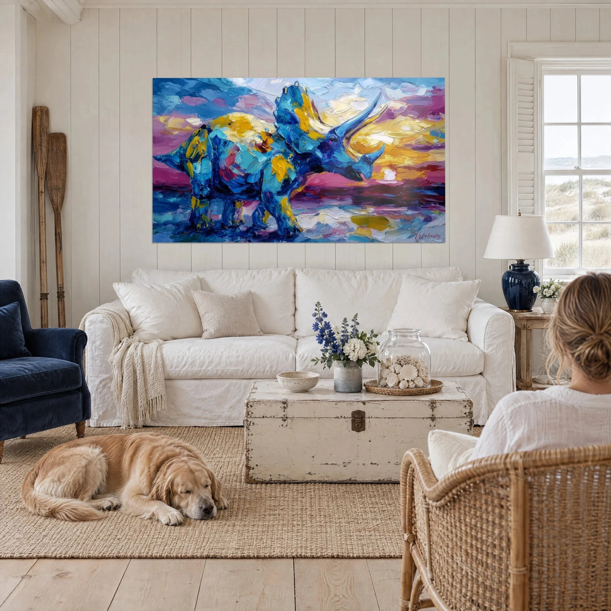 Wall art blue triceratops with golden crest against a violet sky, a woman seen from behind and her dog, above a sofa in a bright living room