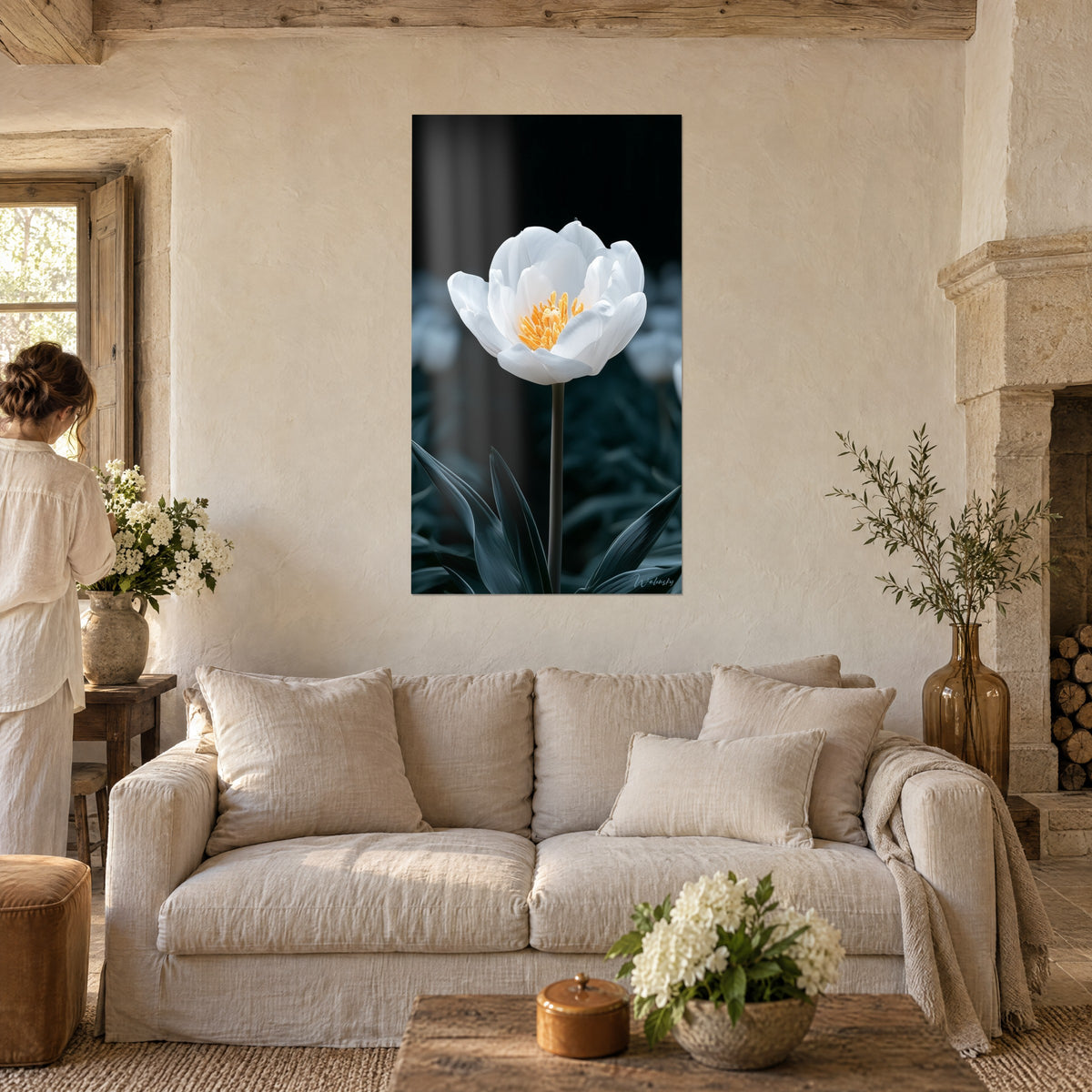 Wall art white tulip with orange stamens on a black background, blurred blue-grey foliage, a woman seen from behind, above a sofa in a bright living room