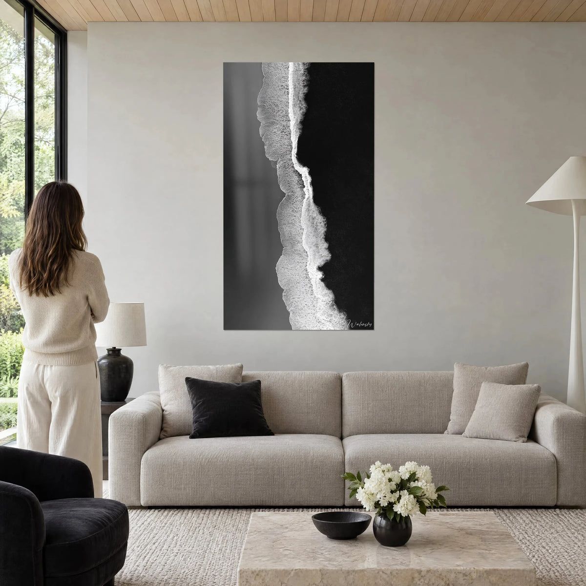 Wall art aerial view of a shoreline, white lacy surf on black sand, woman seen from behind, above a sofa in a bright living room