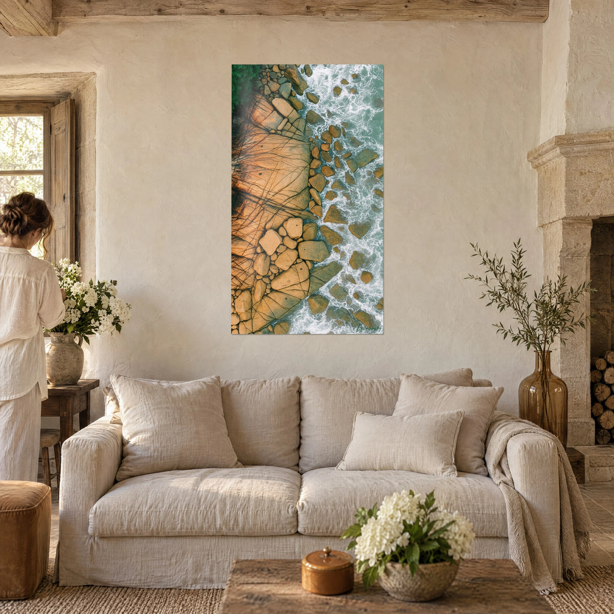 Gemälde ockerfarbene Felsen mit Rissen, gesäumt von weißer Gischt und jadegrünem Wasser, eine Frau von hinten, über einem Sofa in einem hellen Wohnzimmer