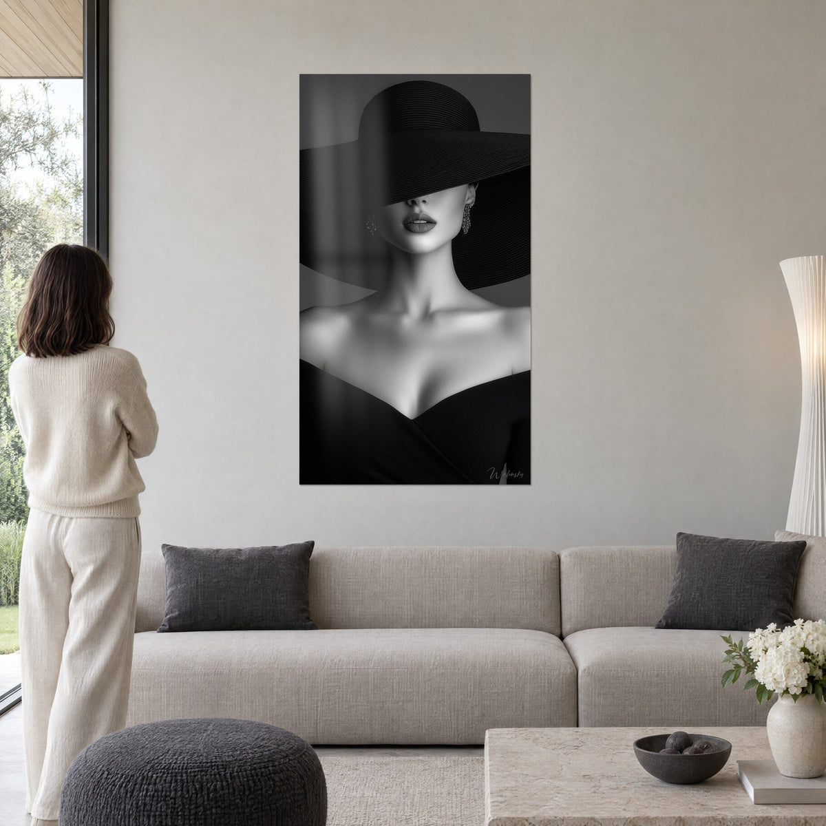 Quadro donna con un enorme cappello nero, labbra scure e spalle nude in bianco e nero, sopra un divano in un soggiorno luminoso