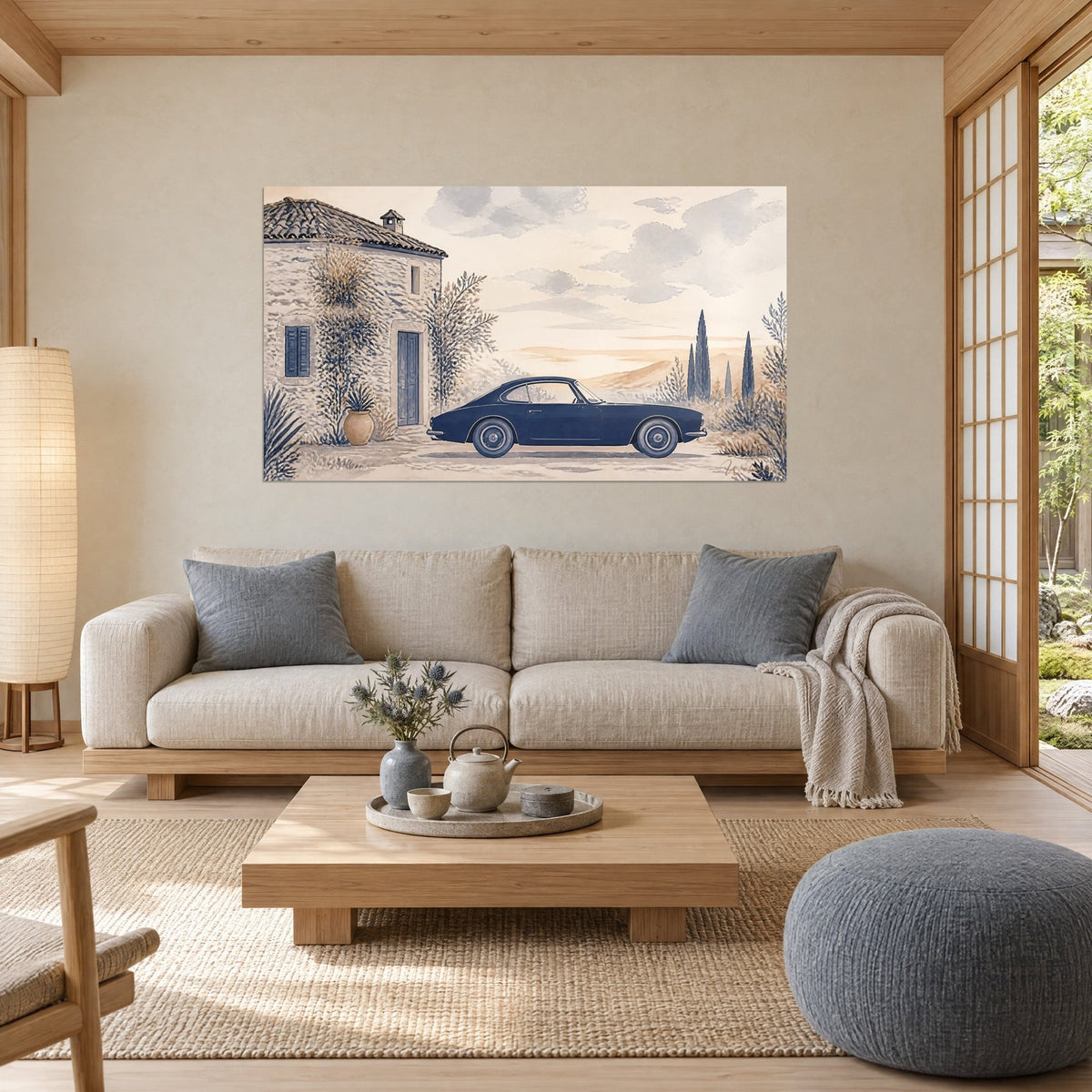 Wall art of a navy blue coupe parked in front of a stone farmhouse, dark cypress trees and cream sky, above a sofa in a bright living room