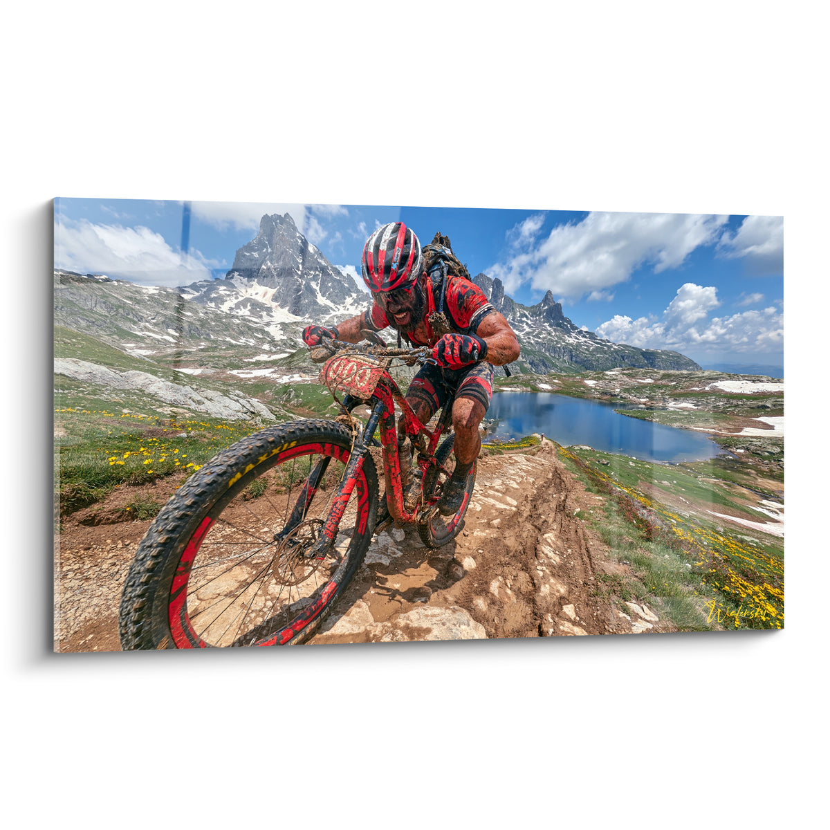 Cycliste en rouge et noir dévalant un sentier rocheux face à des sommets enneigés et un lac bleu