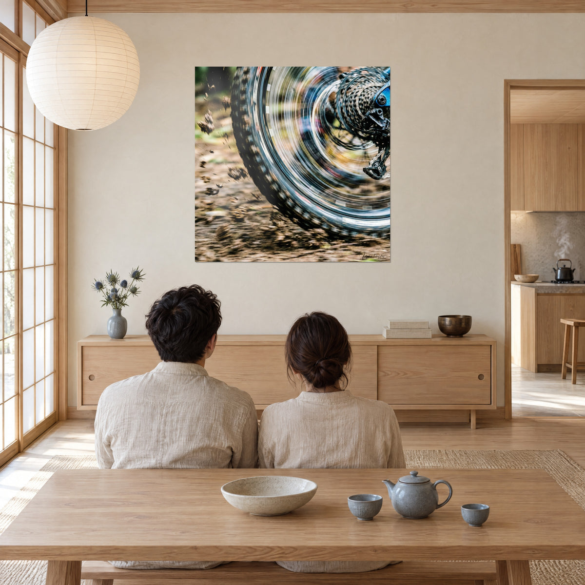 Wall art of a forest trail seen through the mountain bike wheel, blurred silver spokes and brown dirt clumps, viewed by a couple from behind in the living room