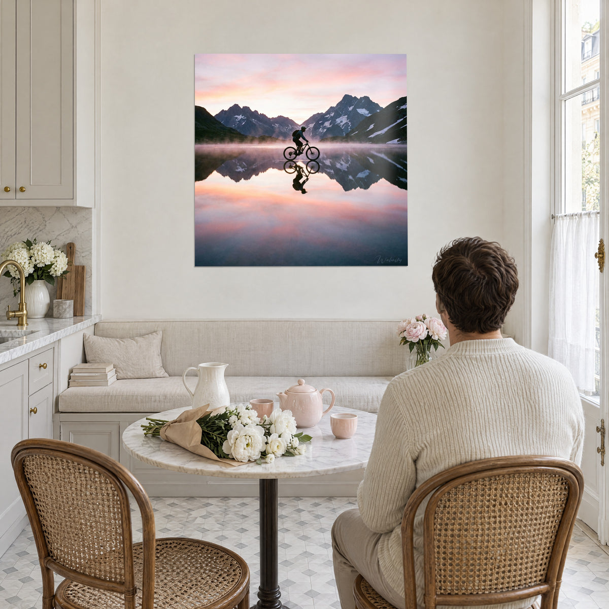 Schilderij ochtendbergen en de spiegeling van een fietser, zalmkleurige lucht, leikleurige besneeuwde toppen, tegenover een man aan de keukentafel
