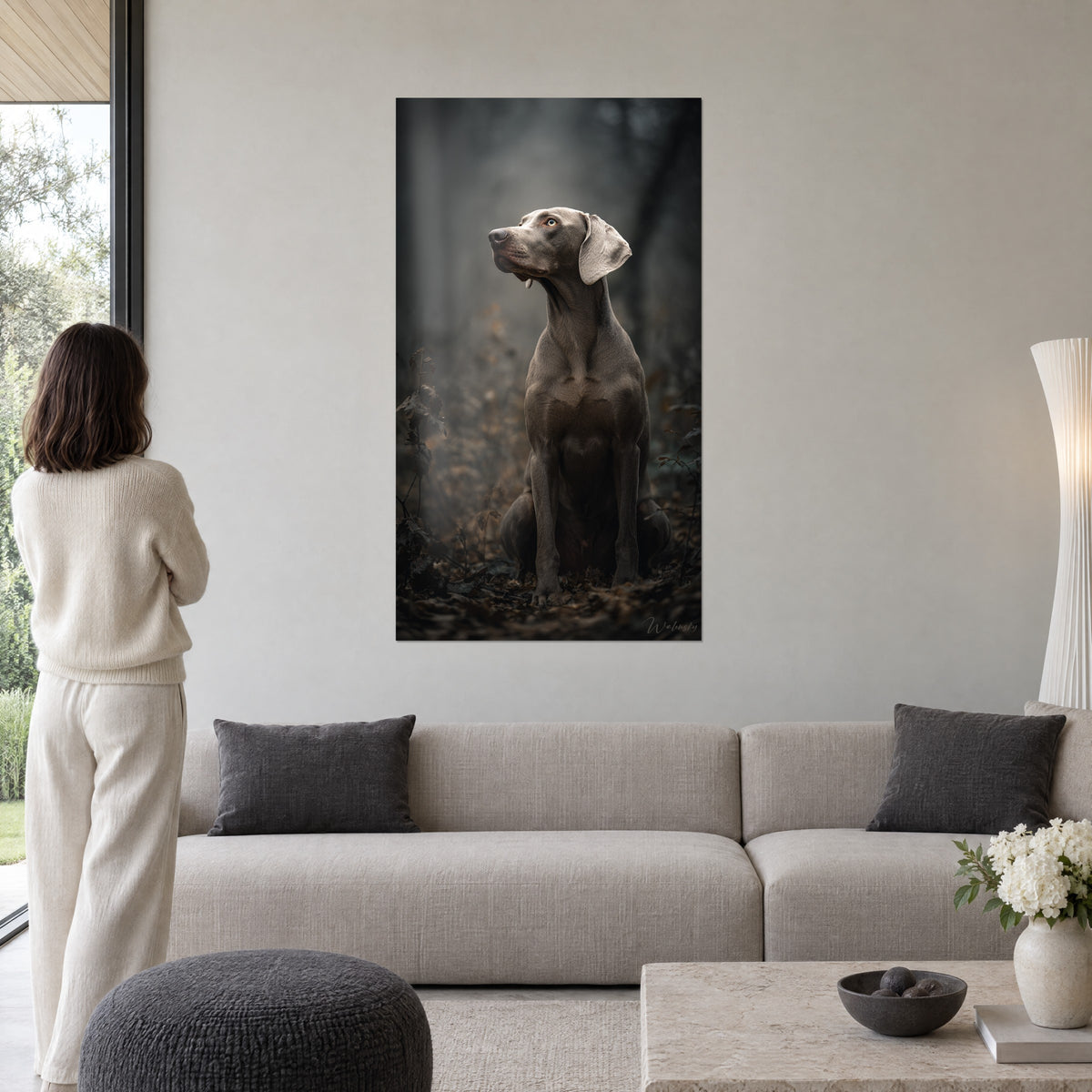 Cuadro de un weimaraner gris sentado con mirada dorada alzada en un bosque brumoso marrón, mujer de espaldas, encima de un sofá en un salón luminoso