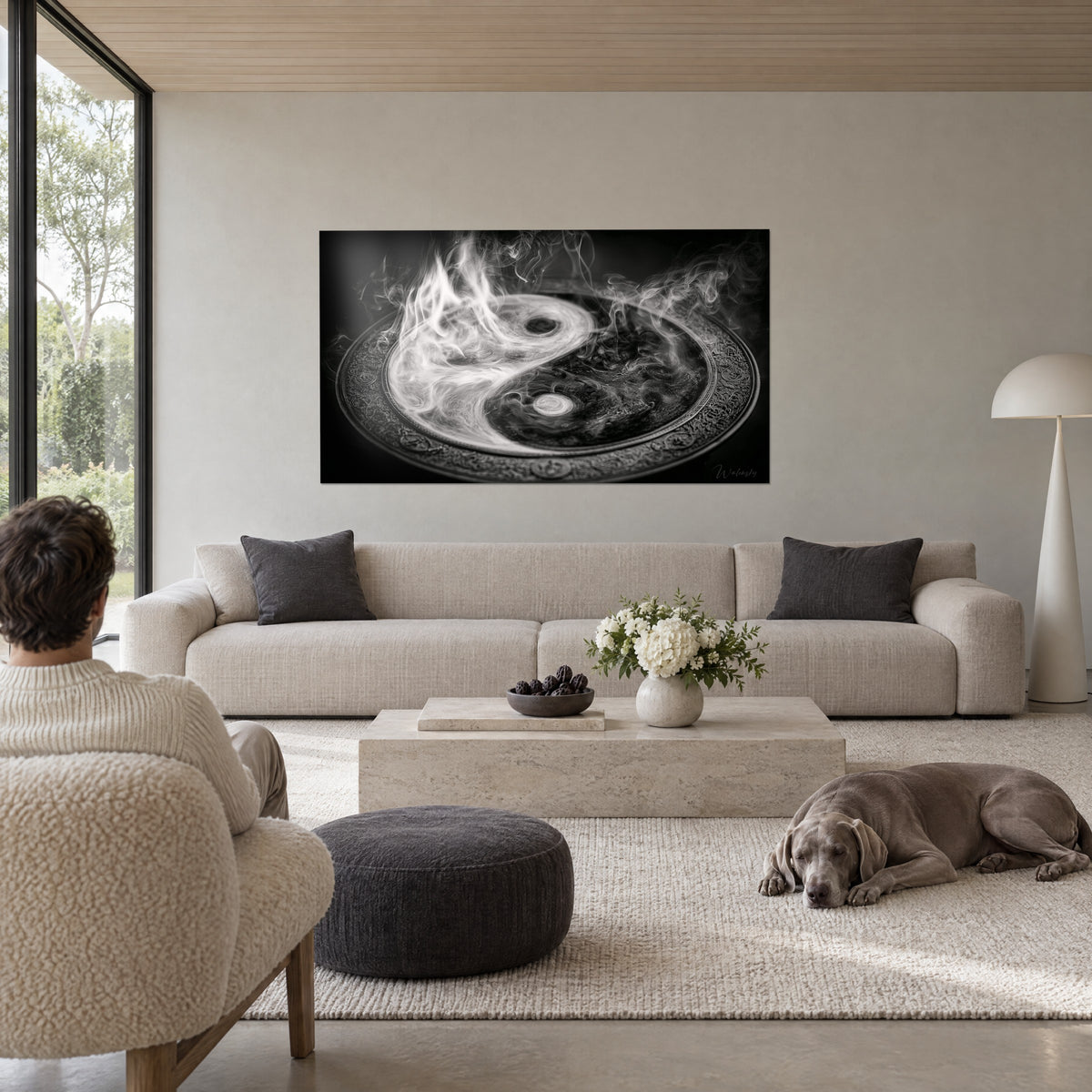Wall art yin yang of white and black smoke on an engraved disc, facing a man seen from behind and a Weimaraner, above a sofa in a bright living room