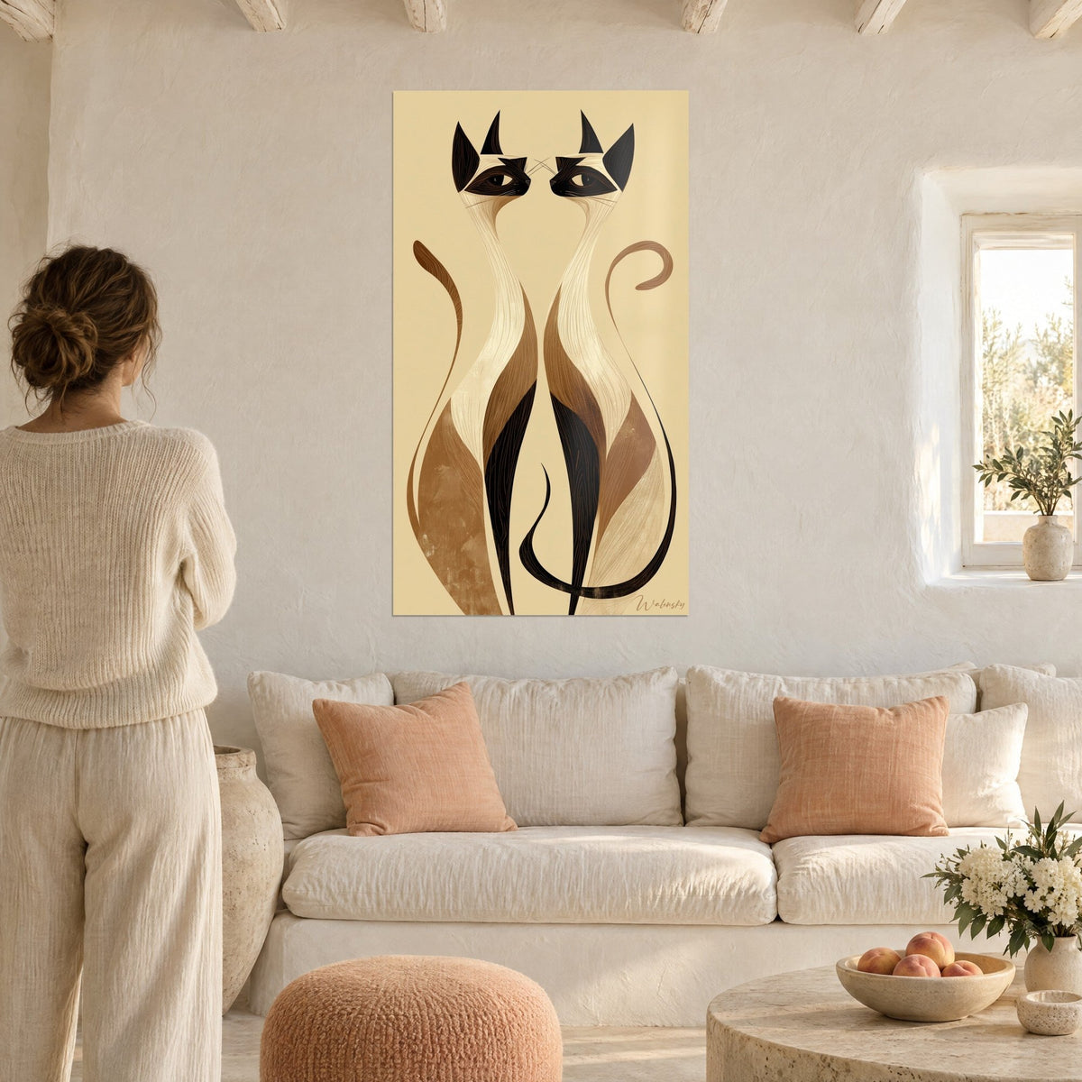 Schilderij twee Siamese katten met langgerekte nekken, zwart en karamel op een zandkleurige achtergrond, vrouw van achteren boven een bank in een lichte woonkamer