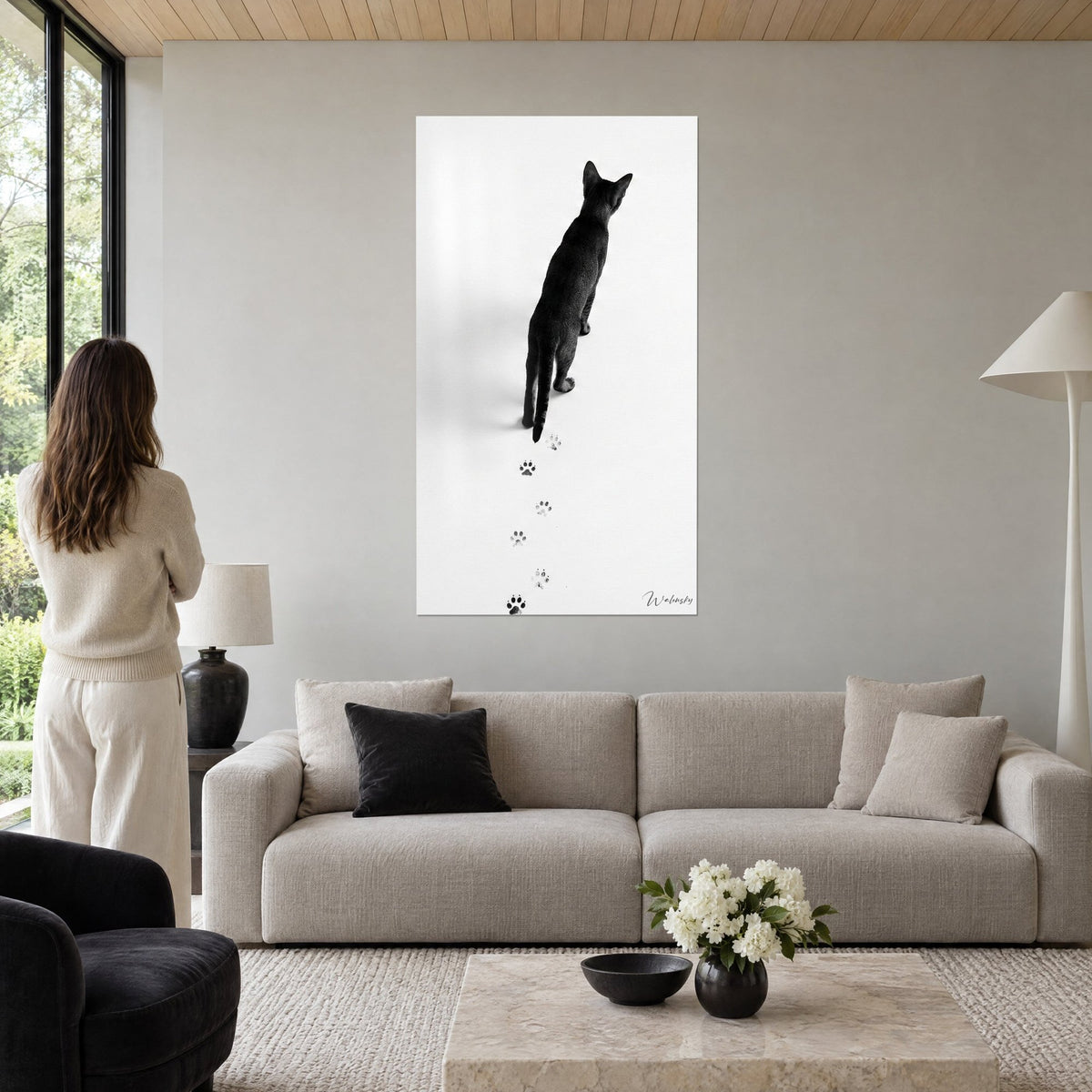 Wall art black cat seen from behind on white background, grey paw prints in a line, vertical frame, above a sofa in a bright living room
