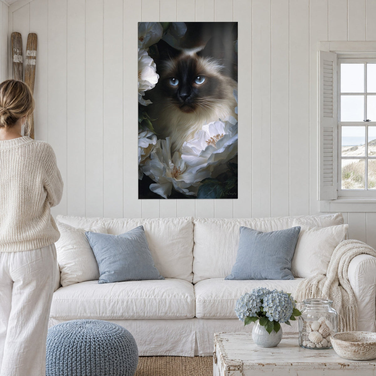 Gemälde Birmakatze mit blauen Augen zwischen weißen Pfingstrosen, cremefarbenes und schokoladenbraunes Fell, über einem Sofa in einem hellen Wohnzimmer