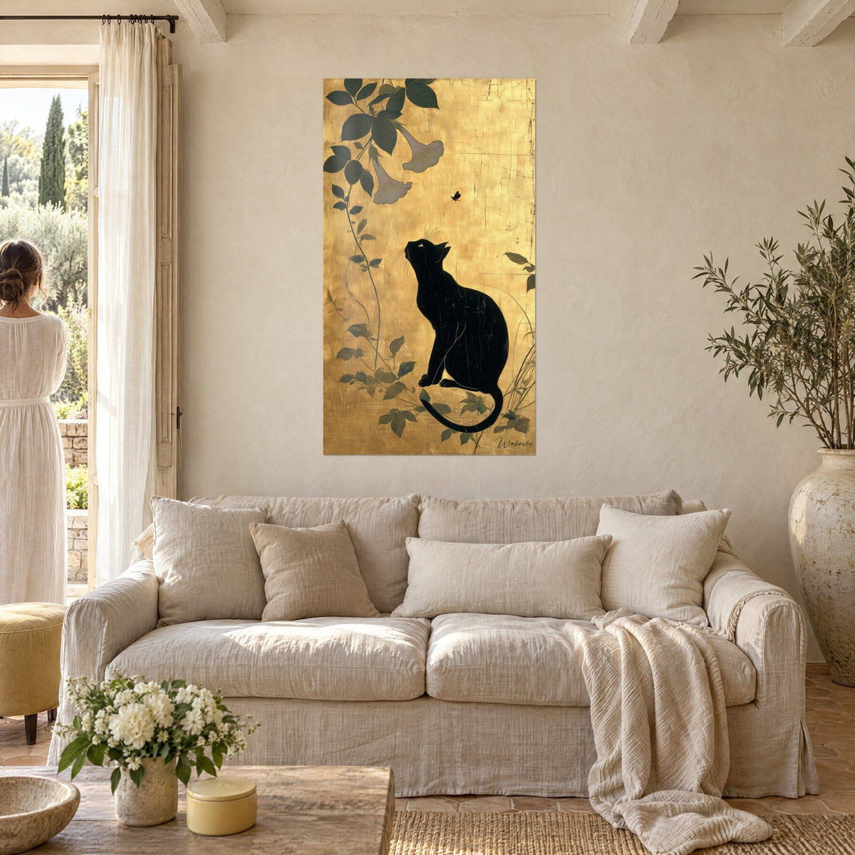 Wall art black cat sitting, muzzle raised toward two white trumpet flowers on cracked gold leaf, above a sofa in a bright living room