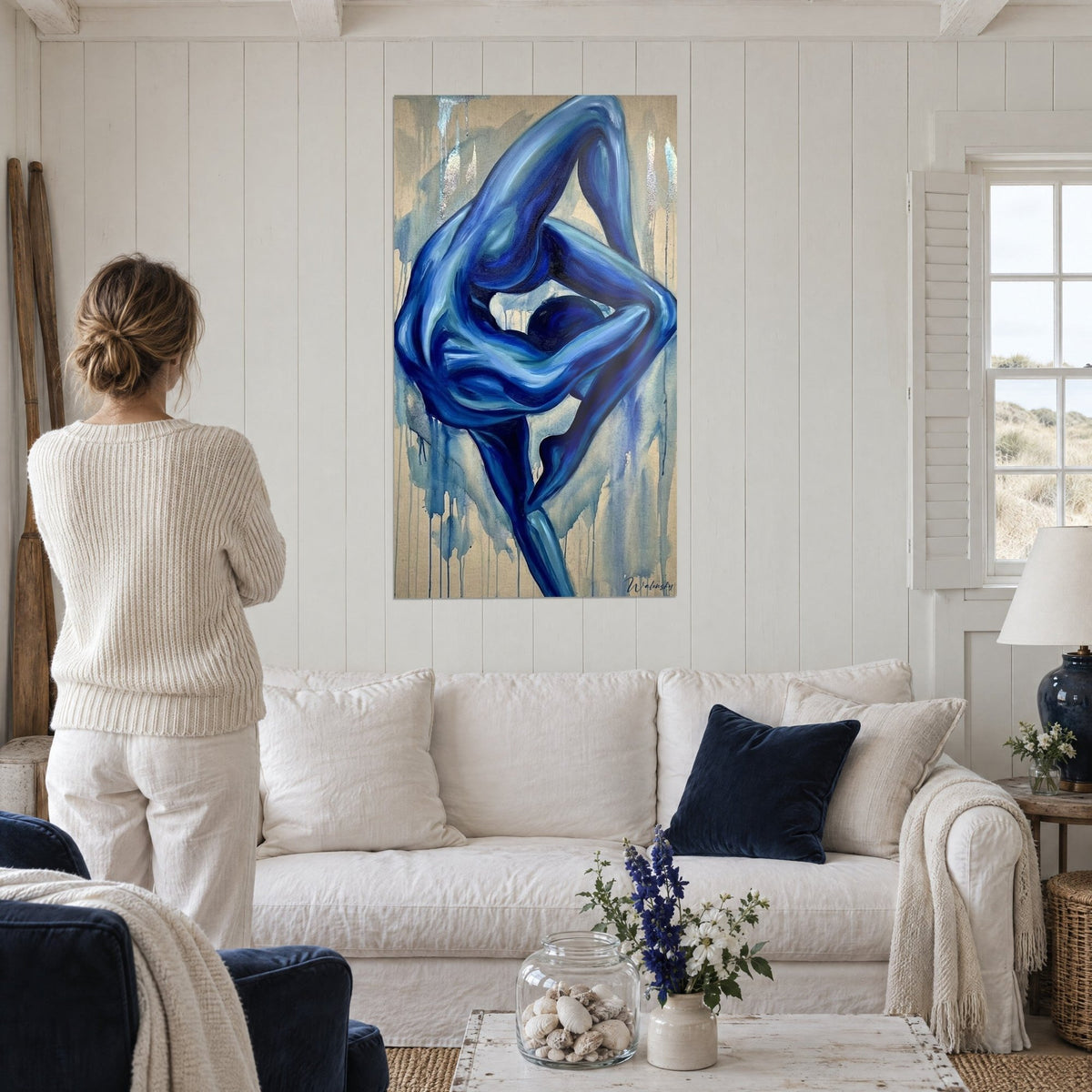 Cuadro contorsionista en azul cobalto, pierna elevada por encima de la cabeza, fondo arena, encima de un sofá en un salón luminoso