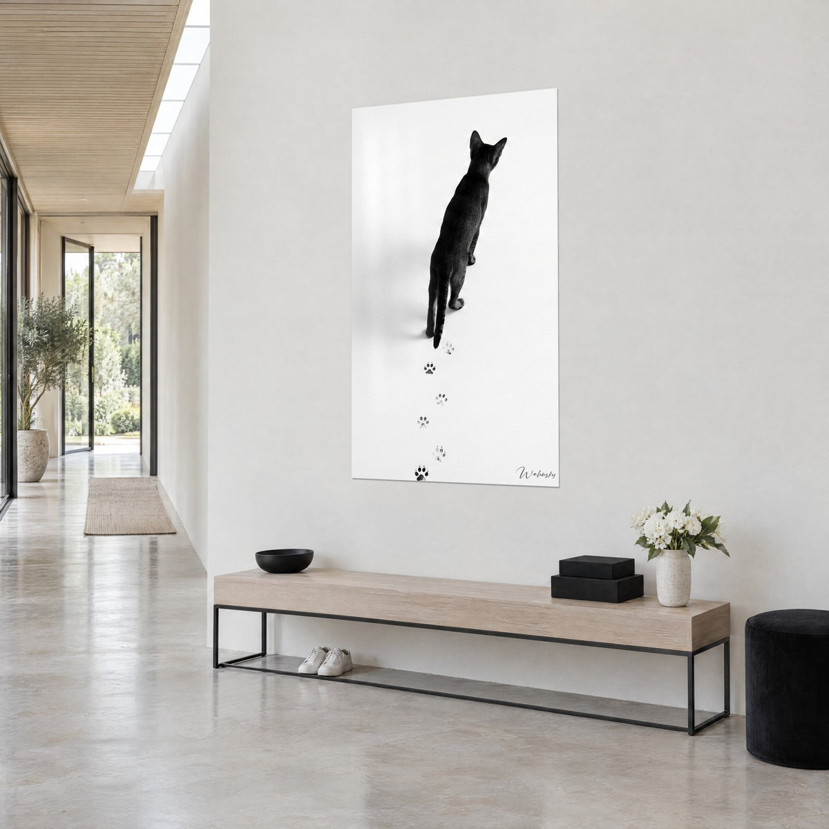 Wall art ebony cat seen from behind, slender silhouette, upright ears and dark grey paw prints, white wall, light wooden bench